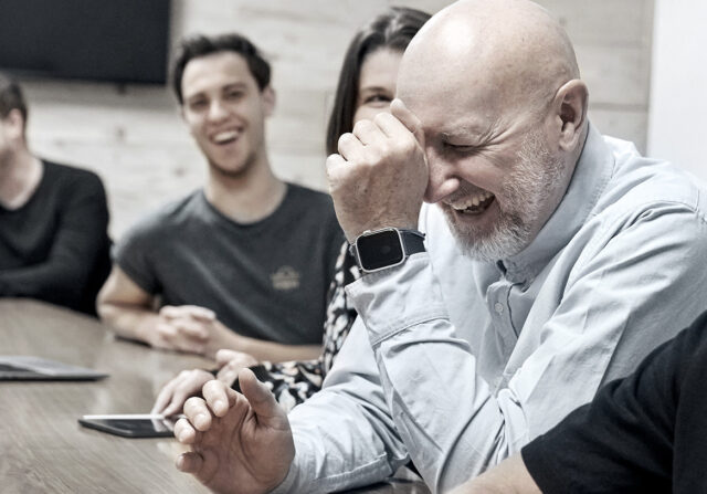 Eine Gruppe von Menschen sitzt um einen Tisch herum und lacht gemeinsam. Der Mann im Vordergrund hält sich die Stirn und lächelt breit. Auf dem Tisch liegen verschiedene Geräte.