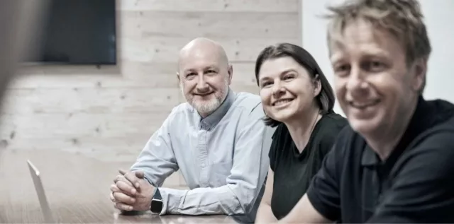 Drei Personen sitzen in einem Konferenzraum an einem Tisch und lächeln in die Kamera.