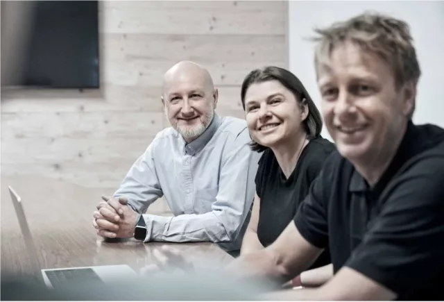 Drei Personen sitzen in einem Konferenzraum an einem Tisch und lächeln in die Kamera.