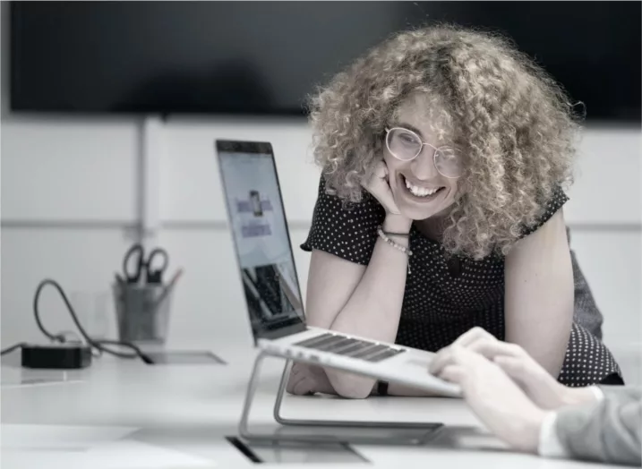 Eine lächelnde Person mit lockigem Haar beugt sich über einen Laptop auf einem Schreibtisch und unterhält sich mit einer anderen Person, deren Hand sichtbar ist. Im Hintergrund sind Büromaterialien zu sehen.