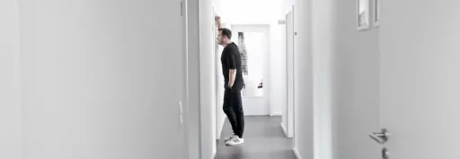 Eine Person, die bei schalk&friends arbeitet, mit schwarzem Hemd und weißen Turnschuhen lehnt in einem schmalen, weißen Flur an einer Wand und blickt ein ein Marketing Büro.